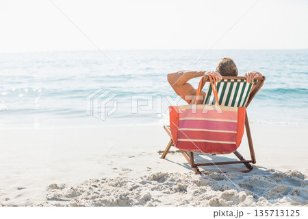 Man relaxing on striped deck chair on sunlit sandy ocean shoreline holding tote bag, copy space 135713125