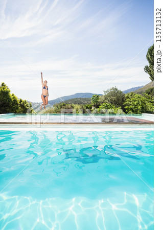 Woman wearing navy bikini jumping from edge of infinity-edge pool, splashing into turquoise water 135713132