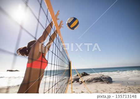 Female athlete in red sportswear reaching to block volleyball over net on beach volleyball court 135713148