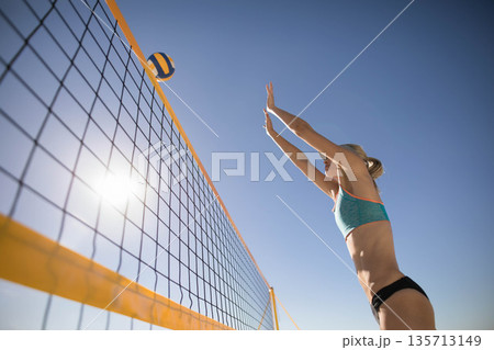 mid-adult athlete leaping in sports bra on beach court blocking ball at volleyball net, copy space mid-adult athlete leaping in sports bra on beach court blocking ball at volleyball net, copy space 135713149
