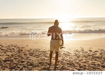 Male surfer standing on sandy shoreline at sunrise wearing board shorts and holding surfboard 135713157