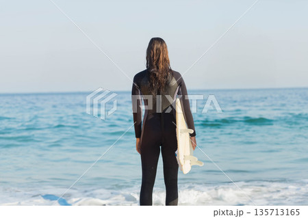 Woman standing at shoreline in wetsuit facing sea while holding white surfboard under right arm 135713165