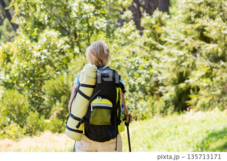Senior woman hiking along sunlit forest trail carrying large backpack and gripping trekking pole 135713171