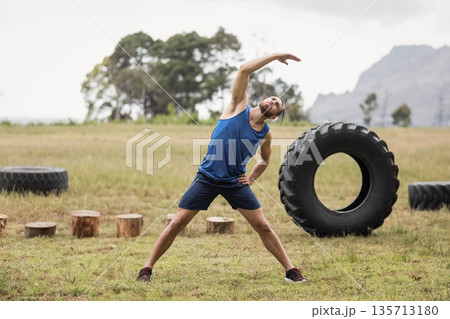 Male fitness enthusiast stretching in open field beside stumps and tractor tires with running shoes Male fitness enthusiast stretching in open field beside stumps and tractor tires with running shoes 135713180