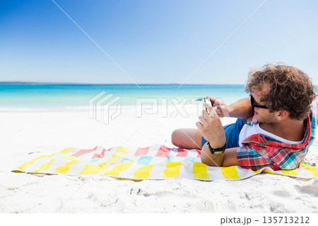 Man lying on towel at beach using smartphone, wearing sunglasses, fitness tracker, copy space 135713212
