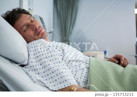 Senior male patient lying in hospital room on bed wearing patterned gown and holding IV line 135713215