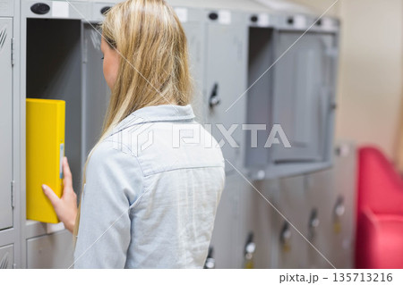Teenage girl wearing denim jacket placing yellow binder into grey locker at school beside red bench 135713216