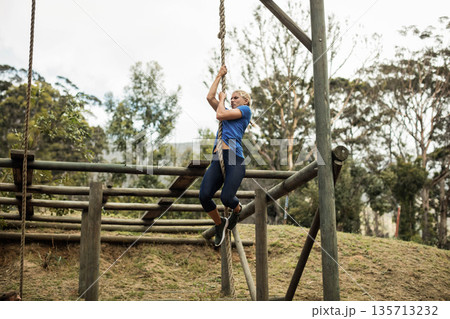 Woman climbing thick rope on obstacle course on grassy slope wearing sportswear and running shoes Woman climbing thick rope on obstacle course on grassy slope wearing sportswear and running shoes 135713232