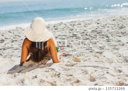 Woman reclining on pale sand near water wearing green bikini bottoms and wide-brimmed white hat Woman reclining on pale sand near water wearing green bikini bottoms and wide-brimmed white hat 135713269