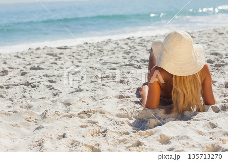 Woman lying on white sand wearing bikini, straw hat gazing toward turquoise waves, copy space 135713270