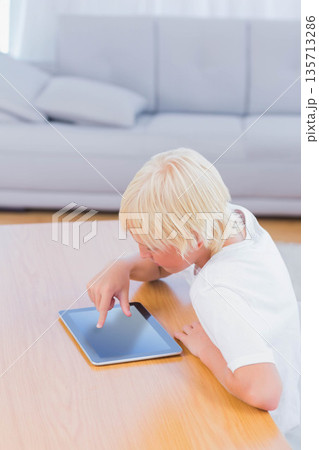 Blond boy tapping tablet screen while leaning on wooden table beside pale sofa in living room Blond boy tapping tablet screen while leaning on wooden table beside pale sofa in living room 135713286