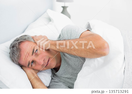 Middle-aged man lying on bed pulling blanket up pressing hands against ears near lamp in bedroom 135713291