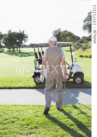 Senior male golfer walking along cart path toward parked golf cart holding club with gloved hand 135713311