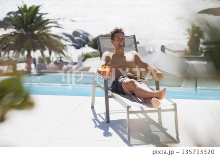 Mid-adult man reclining on metal lounge chair at poolside holding bright orange tropical cocktail 135713320
