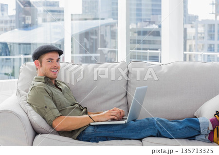 Male reclining on light gray sofa in modern lounge typing on silver laptop, copy space Male reclining on light gray sofa in modern lounge typing on silver laptop, copy space 135713325