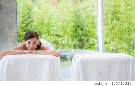 Female spa client lying prone on towel-covered massage table near glass wall with green shrubbery Female spa client lying prone on towel-covered massage table near glass wall with green shrubbery 135713332