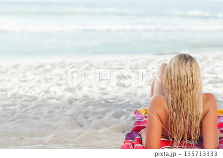 Adult female lying on stomach on striped towel at shore, wearing two-piece swimsuit, copy space 135713333