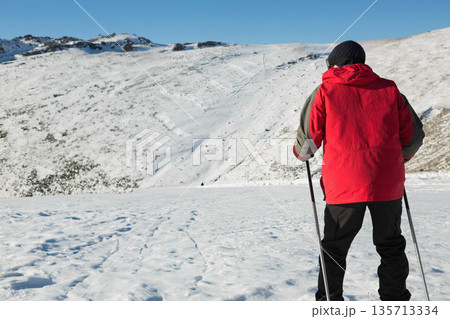 Man wearing ski jacket standing with poles on snowy slope facing blue sky, copy space Man wearing ski jacket standing with poles on snowy slope facing blue sky, copy space 135713334