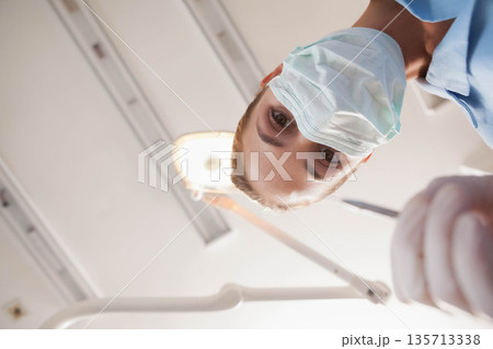 Female dentist wearing mask holding probe in clinic under overhead lamp and ceiling panels Female dentist wearing mask holding probe in clinic under overhead lamp and ceiling panels 135713338