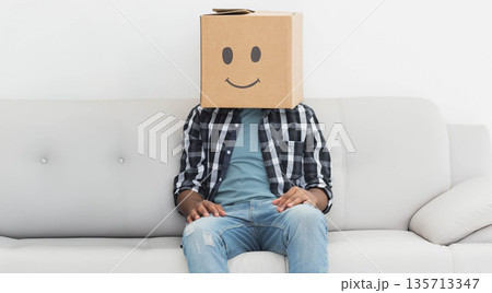 Adult male sitting on light grey sofa against white wall, wearing plaid shirt, cardboard box mask Adult male sitting on light grey sofa against white wall, wearing plaid shirt, cardboard box mask 135713347