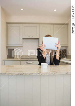 Businesswoman sitting at home behind kitchen island holding tablet computer obscuring face with mug Businesswoman sitting at home behind kitchen island holding tablet computer obscuring face with mug 135713349