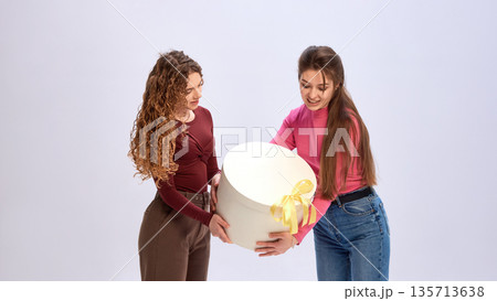 Two female friends holding large round gift box together in studio. Two female friends holding large round gift box together in studio. 135713638