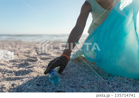 African American woman with black glove collecting plastic water bottle on beach, copy space African American woman with black glove collecting plastic water bottle on beach, copy space 135714741