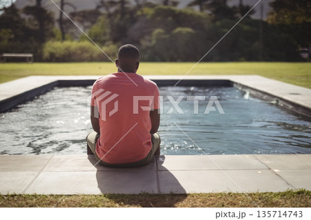 Mid adult African American man sitting on poolside patio in backyard viewing rippling pool water 135714743