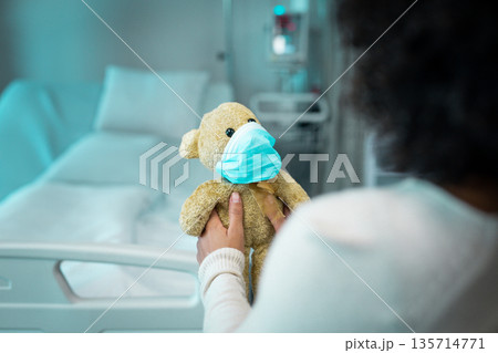 African American woman holding teddy bear wearing blue mask with hospital bed monitor and IV stand African American woman holding teddy bear wearing blue mask with hospital bed monitor and IV stand 135714771