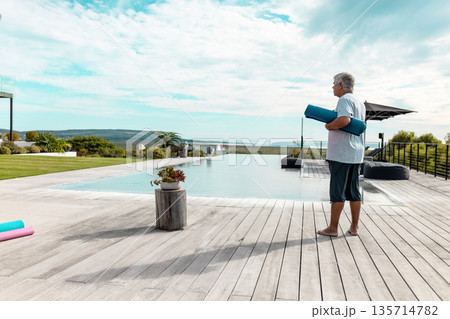 Senior man standing barefoot on wooden deck beside infinity pool holding teal yoga mat, copy space Senior man standing barefoot on wooden deck beside infinity pool holding teal yoga mat, copy space 135714782