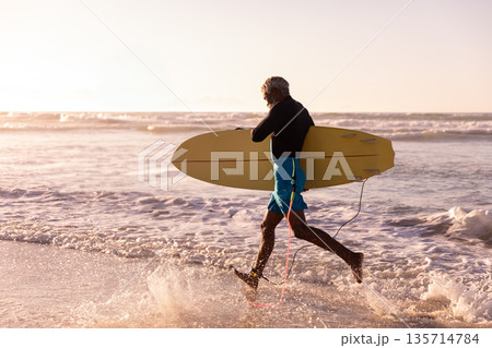 Senior African American man in swim shorts carrying yellow surfboard wading on beach at sunset 135714784