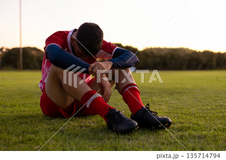 Adult male soccer athlete sitting on grass turf at dusk wearing red jersey and black cleats 135714794