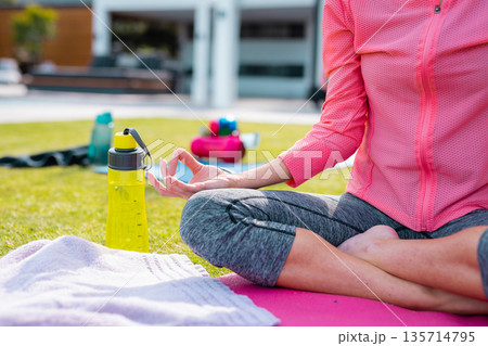 Woman meditating on yoga mat on lawn, forming mudra near towel and water bottles beside dumbbells 135714795