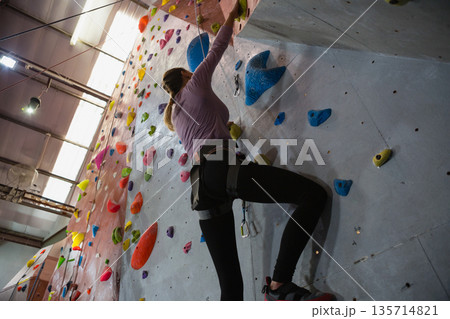 Female climber ascending climbing wall in gym with holds under lights and beams using harness, rope 135714821