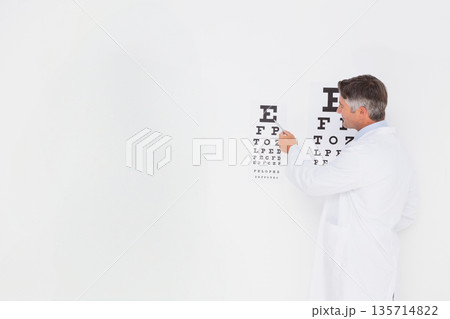 Senior male optometrist pointing with pointer at Snellen eye chart in eye exam room, copy space 135714822