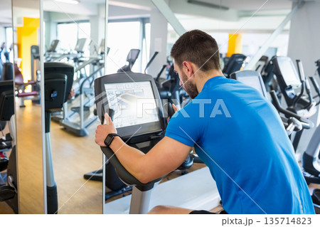 Male gym-goer pedaling on exercise bike console in gym with mirrors and large windows, copy space Male gym-goer pedaling on exercise bike console in gym with mirrors and large windows, copy space 135714823