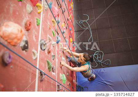 Woman climbing steep red wall at climbing gym using colorful holds and wearing harness, copy space 135714827
