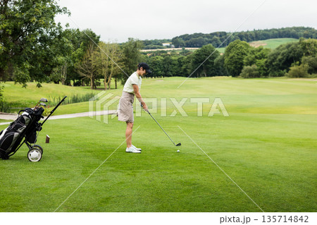 Senior female golfer standing on fairway addressing golf ball with club and push cart, copy space Senior female golfer standing on fairway addressing golf ball with club and push cart, copy space 135714842
