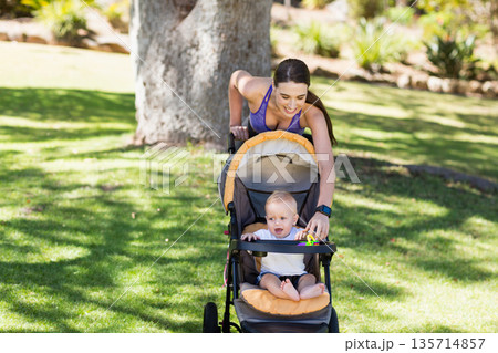Mother and infant son leaning over stroller playing with colorful ring toy on sunlit park lawn 135714857