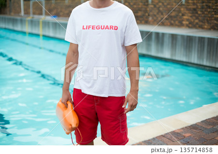 Lifeguard standing at pool edge holding orange rescue buoy beside turquoise water and brick deck 135714858