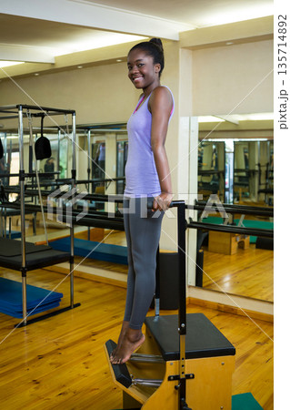 African American woman wearing purple activewear standing on pilates chair in studio rising on toes 135714892