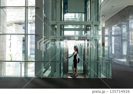 Female professional walking past glass elevator shaft inside corporate lobby with bag 135714916
