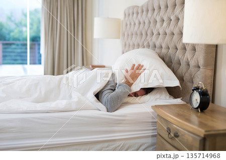 Man lying under white duvet in bedroom covering head with pillow on bed beside nightstand lamp Man lying under white duvet in bedroom covering head with pillow on bed beside nightstand lamp 135714968