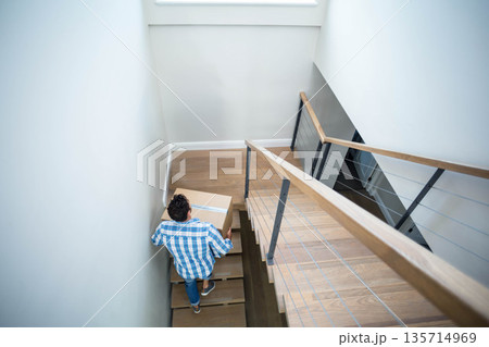 Man carrying sealed cardboard moving box ascending wooden staircase in modern home Man carrying sealed cardboard moving box ascending wooden staircase in modern home 135714969