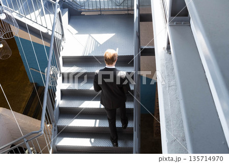 Metal staircase rising in industrial stairwell, sunlight casting geometric shadows on steel steps Metal staircase rising in industrial stairwell, sunlight casting geometric shadows on steel steps 135714970