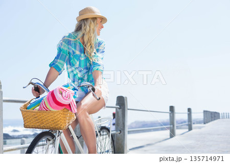 Woman wearing straw hat sitting on bicycle on pier with basket of pastel towels, copy space Woman wearing straw hat sitting on bicycle on pier with basket of pastel towels, copy space 135714971