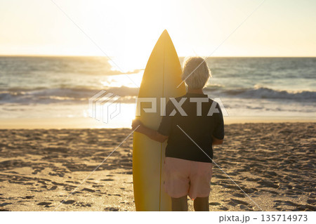 Senior woman standing on sandy shoreline at sunset holding yellow surfboard upright by ocean Senior woman standing on sandy shoreline at sunset holding yellow surfboard upright by ocean 135714973