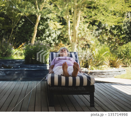 Senior woman relaxing on lounge chair on deck in backyard garden beside reflective water feature Senior woman relaxing on lounge chair on deck in backyard garden beside reflective water feature 135714998