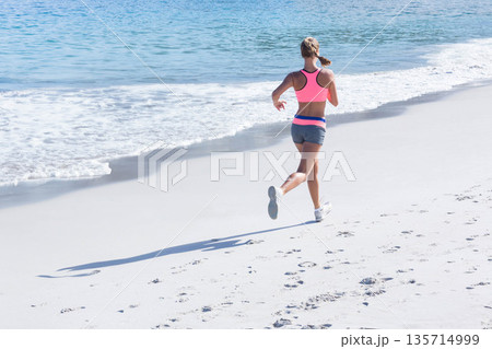 Woman jogging on sandy beach in pink bra, gray shorts, white shoes by ocean, copy space 135714999