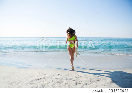 Woman walking on sandy beach carrying surfboard while wearing green swimsuit toward calm blue ocean Woman walking on sandy beach carrying surfboard while wearing green swimsuit toward calm blue ocean 135715031